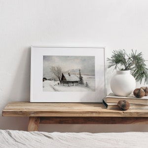 Op de afbeelding: Een ingelijst aquarel schilderij van een besneeuwde winterse sc&egrave;ne met een klein huisje en een boom op de voorgrond. Het schilderij is in een wit lijst en staat op een houten plank met een vaas en decoratieve ornamenten.