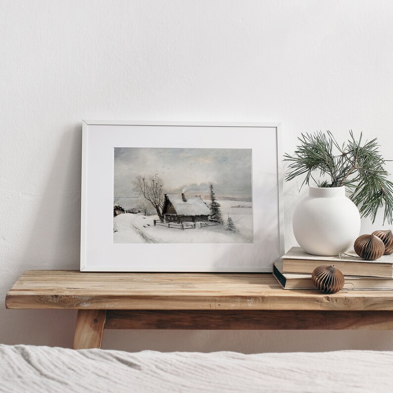 Op de afbeelding: Een ingelijst aquarel schilderij van een besneeuwde winterse sc&egrave;ne met een klein huisje en een boom op de voorgrond. Het schilderij is in een wit lijst en staat op een houten plank met een vaas en decoratieve ornamenten.