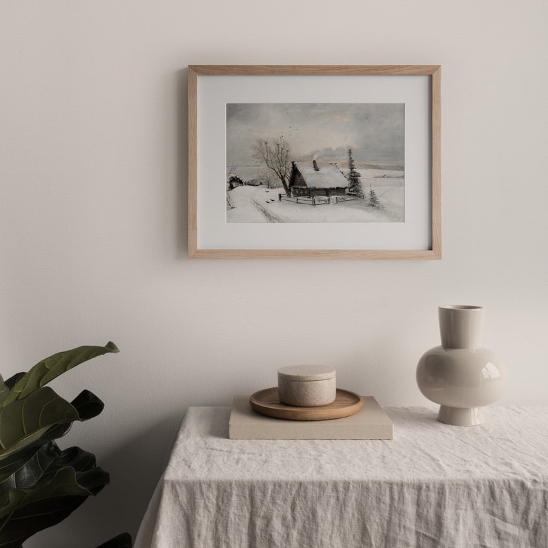 Op de afbeelding: Een ingelijst aquarel schilderij van een klein huisje in een besneeuwd landschap. Het huisje heeft een bruin dak en een witte schoorsteen. Er staat een boom voor het huisje en een pad leidt er naartoe. De lucht is lichtblauw met wat wolken.