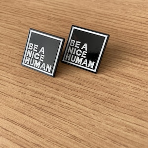 May include: Two black enamel pins with white outlines and text. The pins say "Be a Nice Human".