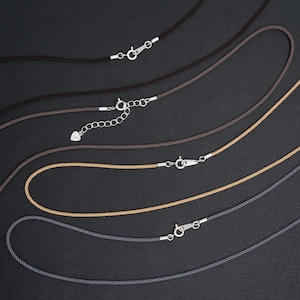 May include: A collection of cord necklaces in various shades of brown, tan, and gray, each with a silver-colored clasp. The necklaces are displayed against a dark background, showcasing their simple design and color variations.