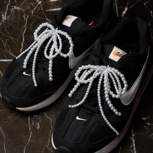 Op de afbeelding: Zwarte sneakers met witte accenten en parelveters die in een strik zijn geknoopt. De veters zijn lang en hangen aan de zijkanten van de schoenen.