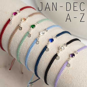 May include: A set of 10 colorful string bracelets with a silver charm and gemstone. Each bracelet has a different color and a letter charm from A to J. The bracelets are arranged in a row on a white surface. The text "JAN - DEC A - Z" is visible in the background.