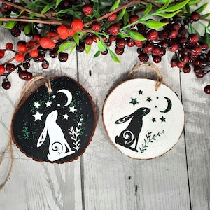 May include: Two wooden ornaments with a black and white hare silhouette design. The hare is looking up at a crescent moon and stars. One ornament is painted black with green glitter, the other is white.