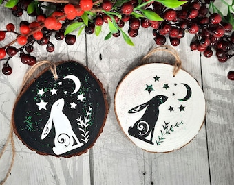 May include: Two wooden ornaments with a black and white hare silhouette design. The hare is looking up at a crescent moon and stars. One ornament is painted black with green glitter, the other is white.