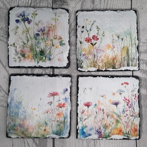 May include: A set of four square slate coasters, each with a unique watercolour floral design. The coasters have a white background and a black border. The floral designs feature colourful wildflowers in shades of pink, purple, yellow, and orange.