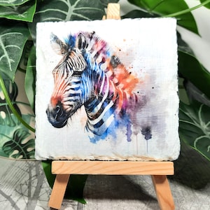 May include: A watercolour painting of a zebra on a small easel. The zebra is painted in shades of blue, purple, and orange. The background is white with watercolour splatters.