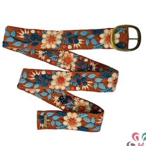 Puede incluir: Cinturón decorativo con estampado floral. El cinturón es de color marrón cálido con flores bordadas en tonos naranja, crema y azul. Tiene una hebilla color latón y una textura tejida.