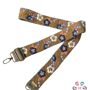 May include: An olive green adjustable bag strap featuring intricate floral embroidery in shades of navy blue, white, and peach. The strap includes antique brass-toned metal hardware, including swivel lobster clasps and a slider, with a small rectangular tag labeled Makantu.