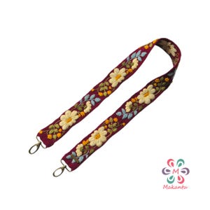May include: A burgundy embroidered bag strap featuring a floral design with cream-colored flowers, blue leaves, and yellow accents. The strap has gold-colored metal clasps on each end for attachment.