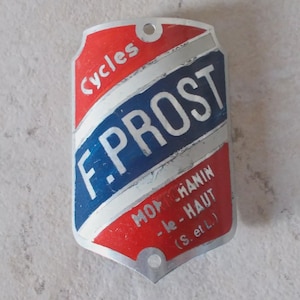 Peut inclure: Plaque de vélo vintage aux couleurs rouge, blanc et bleu. La plaque porte l'inscription "Cycles F. PROST" en blanc, avec du texte supplémentaire en rouge. La plaque comporte deux trous de fixation.