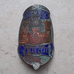 May include: An oval-shaped metal badge featuring a weathered patina with a central embossed design of a castle with two towers. The badge includes blue enamel banners with the text "am.cours" at the top and "sourania" at the bottom in script.