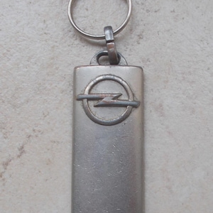 Könnte beinhalten: Ein rechteckiger Schlüsselanhänger aus silberfarbenem Metall mit dem Opel-Logo, das aus einem Kreis mit einem horizontalen Blitz besteht. Der Anhänger hat eine glatte, leicht abgenutzte Oberfläche und einen runden Metallring.