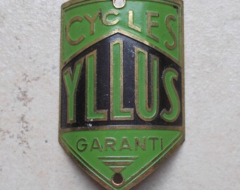 May include: A vintage metal bicycle head badge featuring a shield shape with a green and black enamel finish. The badge displays the text CYCLES YLLUS GARANTI in raised gold-toned lettering, with two small circular holes at the top and bottom.