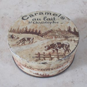 Könnte beinhalten: Eine runde, alte Karamell-Dose mit einer ländlichen Szene. Der Deckel zeigt eine braun-weiße Illustration von Kühen, die in der Nähe eines Holzzauns und eines Chalets grasen. Der Text "Caramels au lait St Christophe" ist auf dem Deckel aufgedruckt.