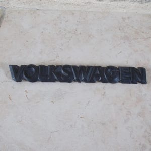 Peut inclure: Emblème VOLKSWAGEN noir en trois dimensions. Les lettres sont stylisées et semblent être faites d'un matériau solide. L'emblème est présenté sur une surface neutre et texturée.