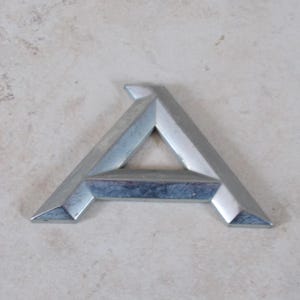 Op de afbeelding: Een zilveren metalen letter "A" met een geometrisch ontwerp. De letter is schuin en heeft een driedimensionale uitstraling. Het oppervlak heeft een geborstelde metaaltextuur, tegen een neutrale achtergrond.