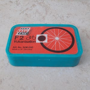 Op de afbeelding: Een rechthoekige opbergdoos van blauwgroen plastic met een rood etiket op het deksel. Het etiket bevat de tekst REMA TIP TOP, F2, Vulcanisation, Art. No. 506 010, Made in W.-Germany, een fietspictogram en een afbeelding van een fietswiel.