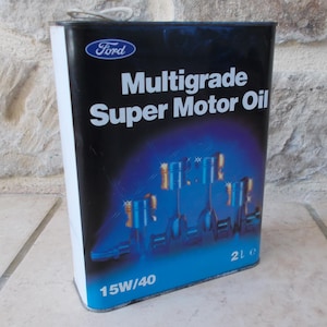Può includere: Un contenitore metallico rettangolare di olio motore Ford Multigrade Super Motor Oil con sfondo sfumato nero e blu. L'etichetta presenta il logo Ford, la scritta 15W/40, un volume di 2L e un'illustrazione grafica di pistoni in blu e oro.