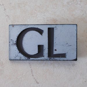 May include: A rectangular, distressed metal sign featuring the letters "GL" in a dark hue. The sign has a light blue-grey background with visible wear, giving it a vintage look. The bold letters are prominent against the background.