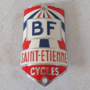 May include: A metal bicycle head badge featuring a red, white, and blue color scheme. The emblem displays the letters BF at the top, with the text SAINT-ETIENNE and CYCLES printed in red and silver lettering below. It has two circular mounting holes.