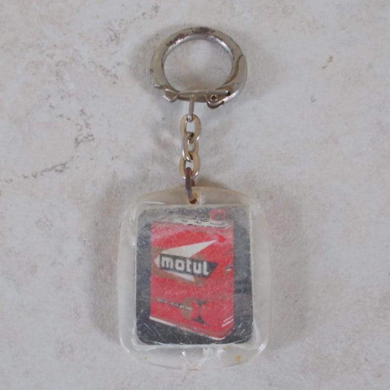 May include: A rectangular clear acrylic keychain featuring an embedded graphic of a red Motul oil can with white and black accents. The keychain is attached to a silver-toned metal split ring by a short chain, showcasing a vintage automotive design.