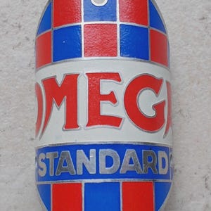 May include: Vintage metal badge with a red, white, and blue color scheme. The badge has a checkered pattern on the top and bottom, with the word "OMEGA" in red letters on a white background, and "STANDARD" in silver letters.