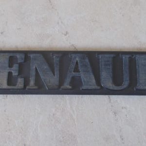 Op de afbeelding: Een rechthoekig, donkergrijs embleem met de naam "RENAULT" in verhoogde, verweerde letters. De letters hebben een lichtere grijstint, wat een contrast creëert met de donkere achtergrond. Het embleem is waarschijnlijk voor een voertuig.