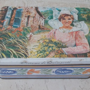 Könnte beinhalten: Rechteckige Blechdose mit einer gemalten Illustration einer Frau in traditioneller bretonischer Tracht, die Blumen hält. Die Dose zeigt eine Szene eines Steingebäudes und florale Akzente, mit dem Text "Costume de Quimperlé".