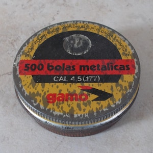May include: A round metal tin with a yellow and black design. The tin has the text "500 bolas metalicas CAL 4.5 (.177)" in red and white. The brand name "gamo" is in red. The tin is for round shots.