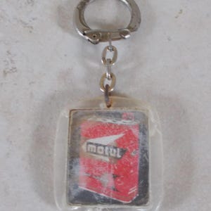 May include: A rectangular clear acrylic keychain featuring an embedded graphic of a red Motul oil can. The keychain includes a silver-toned metal split ring attached by a short chain, showcasing a vintage-style design with the Motul brand logo.