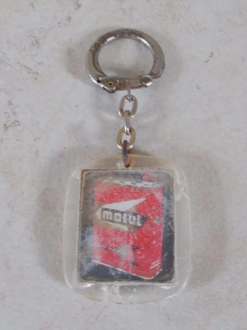 May include: A rectangular clear acrylic keychain featuring an embedded graphic of a red Motul oil can. The keychain includes a silver-toned metal split ring attached by a short chain, showcasing a vintage-style design with the Motul brand logo.