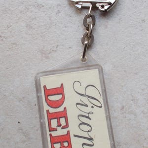 Peut inclure: Un porte-clés rectangulaire transparent avec un anneau argenté. Le porte-clés contient un insert crème avec le nom "Deb" en lettres rouges et "Firon" en cursive noire. Il est attaché à une chaîne argentée.