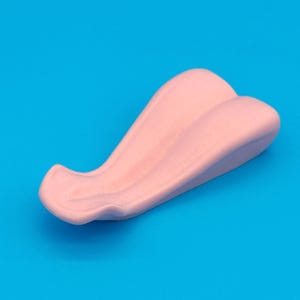 Puede incluir: Un objeto curvo de color rosa claro con una superficie lisa, sobre un fondo azul brillante. El objeto tiene una forma redondeada con una ligera hendidura en un lado. El logotipo "FURRY FACTORY" está en la parte inferior.