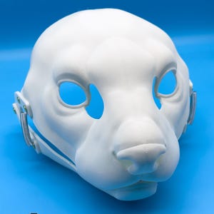 Peut inclure: Un masque animalier blanc imprimé en 3D, avec de grandes ouvertures pour les yeux et un museau détaillé. Le masque comporte des charnières et des ressorts métalliques sur les côtés. Le logo "FURRY FACTORY" est visible en bas sur fond bleu.