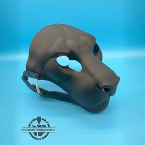 Puede incluir: Una máscara de animal impresa en 3D de color negro mate con un hocico felino, aberturas para los ojos y un mecanismo de mandíbula articulada con un resorte metálico visible en el lateral. El logo FURRY FACTORY aparece impreso abajo a la izquierda.