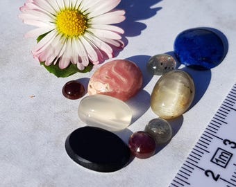 May include: Assortment of eight polished gemstones, including a pink and white stone, a clear stone, a black stone, a blue stone, a gray stone, and two red stones. The stones are arranged on a white surface with a ruler in the background.