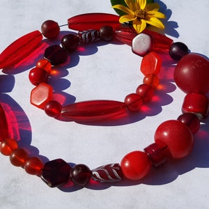 Peut inclure: Un collier de perles en verre rouge, orange et bordeaux. Les perles sont de formes et de tailles variées, notamment rondes, ovales et hexagonales.