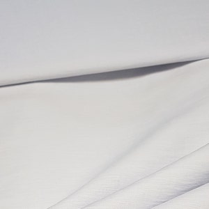 Pode incluir: Tecido branco com uma superfície lisa e ligeiramente texturizada. O tecido está dobrado e drapeado, mostrando sua natureza suave e fluida.