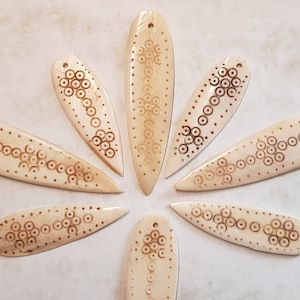 Op de afbeelding: Zeven been hangers met een gesneden ontwerp van cirkels en stippen. De hangers zijn licht crème van kleur en hebben een puntige punt.