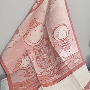 Op de afbeelding: Een roze en witte keukendoek met een grillige illustratie van een gezin. Het ontwerp toont een moeder, een dochter en een zoon, allemaal lachend. De handdoek heeft een decoratieve rand met harten en een dambordpatroon.