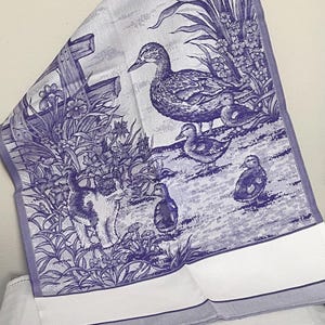 Op de afbeelding: Een blauw-witte keukendoek met een gedetailleerde illustratie van eenden, eendenkuikens en een kat in een tuin. De doek heeft een witte rand en toont een hek en bloemen.