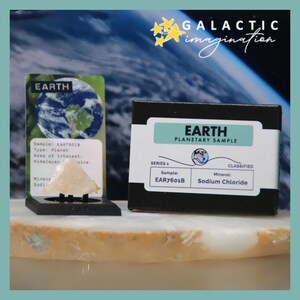 Puede incluir: Una exhibición con una pequeña muestra mineral de color claro etiquetada como "EARTH" con una tarjeta que muestra la imagen de la Tierra. También hay una caja negra con "EARTH PLANETARY SAMPLE", con el texto "Sodium Chloride". El fondo tiene un tema espacial.