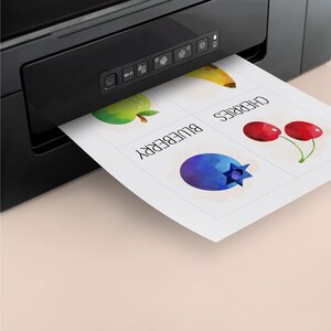 May include: A black printer with a white sheet of paper coming out of it. The paper has four squares with watercolor images of a banana, apple, blueberry, and cherries. The squares are labeled "banana", "apple", "blueberry", and "cherries".