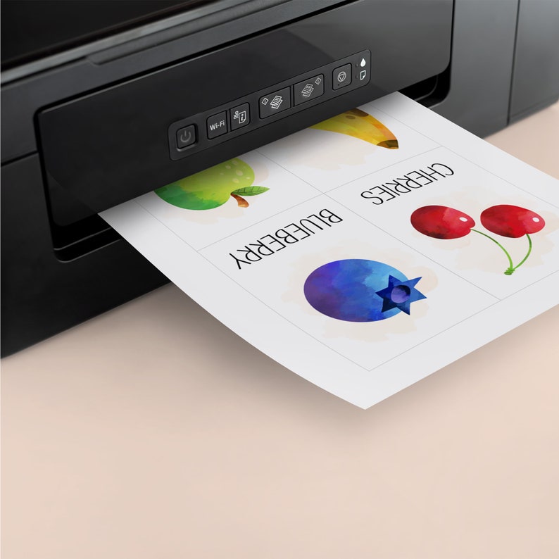 May include: A black printer with a white sheet of paper coming out of it. The paper has four squares with watercolor images of a banana, apple, blueberry, and cherries. The squares are labeled "banana", "apple", "blueberry", and "cherries".