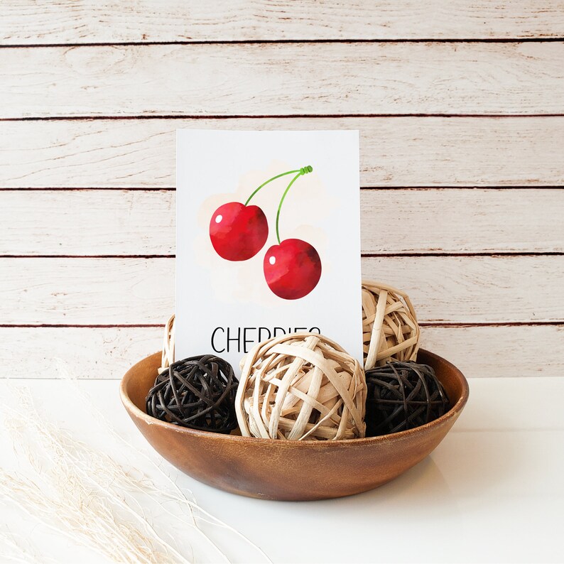 May include: A white card with a watercolor illustration of two red cherries with green stems. The card says "CHERRIES" in black text. The card is sitting on a wooden bowl with brown and beige balls.
