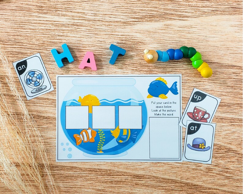 K&ouml;nnte beinhalten: Ein farbenfrohes Lernspiel f&uuml;r Kinder mit einem Aquarium mit drei leeren Feldern und Karten mit Buchstaben und Bildern. Die Karten sagen "an", "up" und "at".