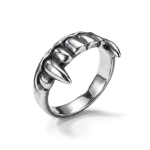 May include: A silver-toned metal ring featuring a sculpted design of multiple pointed teeth or fangs along the top band. The surface has a polished finish with darkened recessed areas to highlight the detailed shape of the individual teeth.