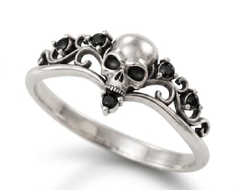May include: A silver ring featuring a central skull motif flanked by intricate scrollwork and five small, round, black faceted stones. The metal has a polished finish with darkened recessed areas to highlight the detailed skull and filigree design.