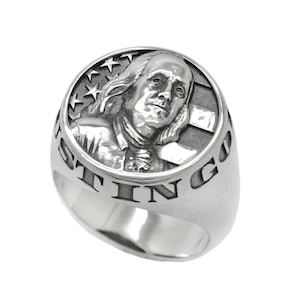 Puede incluir: Un anillo de plata con una imagen en relieve de Benjamin Franklin y la bandera estadounidense. Las palabras "In God We Trust" están grabadas alrededor de la banda.